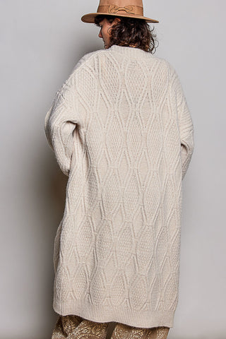 A person shown from the back wearing a chunky cream cable-knit cardigan and a tan hat against a plain light gray studio background.