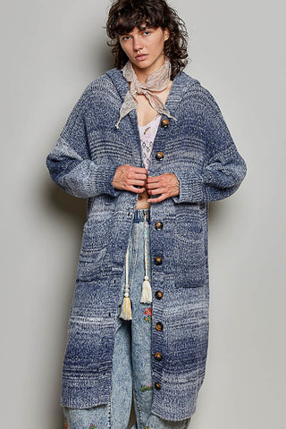 A model wearing a long, oversized blue marled knit cardigan with brown buttons, layered over a patterned top and jeans, against a plain gray studio background.