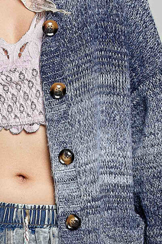 Close-up of a person wearing a marled blue-gray knit cardigan with large dark brown buttons, layered over a pale pink lace top and paired with light-wash denim.