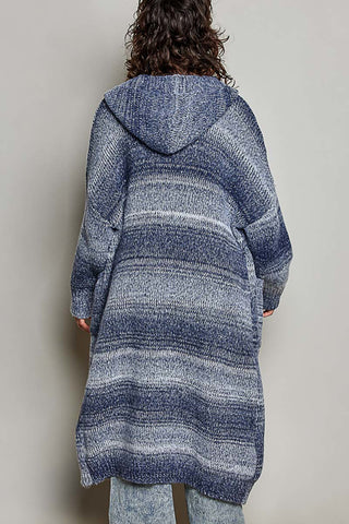 A person shown from the back wearing a long, loose hooded knit cardigan with horizontal blue, gray, and white stripes, paired with light blue jeans, against a plain light gray wall.