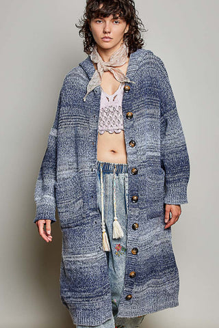 A model wearing a long, oversized blue-gray marled knit cardigan with large brown buttons, layered over a white crochet top and patterned jeans, against a plain gray studio background.