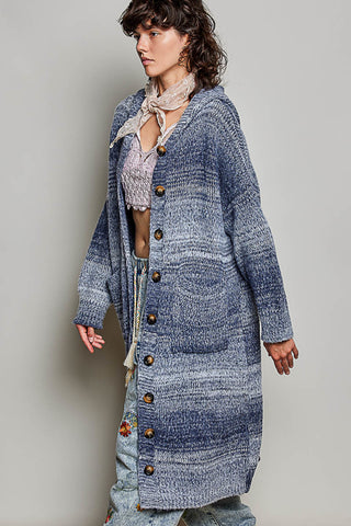A woman models a long, open-front cardigan with large brown buttons, featuring a blue and gray marled knit texture. She is styled with a light pink knit scarf and a light purple cropped top, set against a plain light gray studio background.
