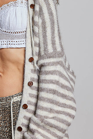 Close-up of a person wearing a fuzzy knit cardigan with horizontal taupe and off-white stripes, brown buttons, and a cropped white lace-trim bandeau top, paired with leopard-print bottoms, against a plain light gray background.