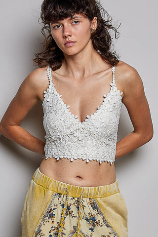 A woman modeling a white lace camisole crop top with floral embroidery and thin straps, paired with yellow patterned pants, against a plain gray studio background.