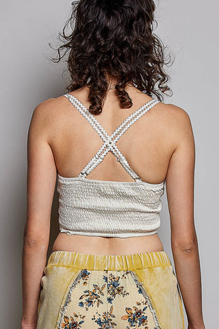 Back view of a woman wearing a textured light beige crop top with braided crisscross shoulder straps, paired with a yellow floral skirt, against a plain gray background.