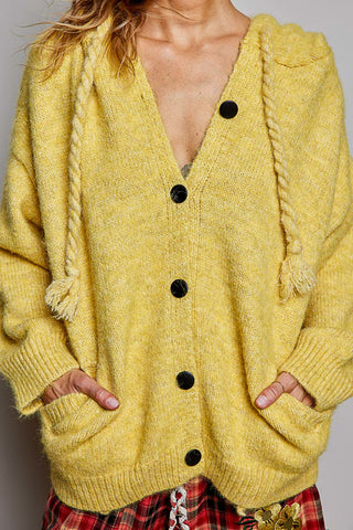 A close-up of a person wearing a chunky knit yellow cardigan with rope drawstrings, black buttons, and front pockets, paired with red plaid bottoms.