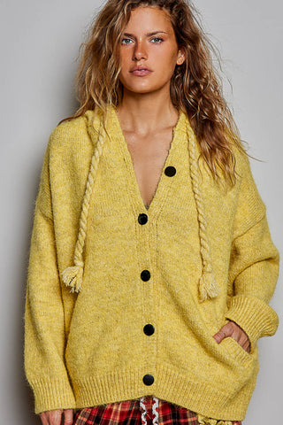 A woman wearing a fuzzy mustard yellow knit cardigan with black buttons and braided drawstrings, paired with a red plaid skirt, against a plain gray studio background.