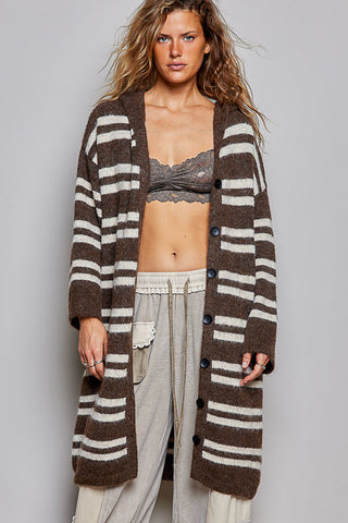 A woman modeling a long brown and cream striped fuzzy knit cardigan with dark buttons, worn open over a lace bralette and light gray pants, against a solid gray studio background.