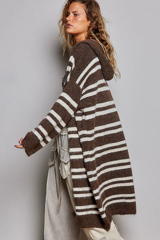 A woman wearing a brown and cream striped fuzzy knit hooded cardigan, paired with light beige trousers, photographed against a solid gray studio background.