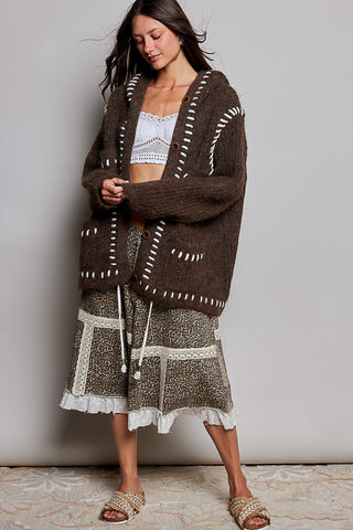 A woman modeling a brown chunky knit cardigan with white stitched details, paired with a brown and white patterned skirt, against a plain gray wall.