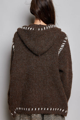 A person viewed from the back wearing a brown textured knit hooded sweater with white contrast stitching details around the hood and shoulders, against a plain light gray background.