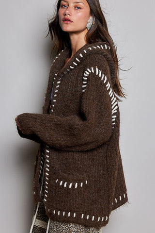 A woman wearing a brown fuzzy knit cardigan with white contrast stitching details, photographed against a plain light gray studio background.