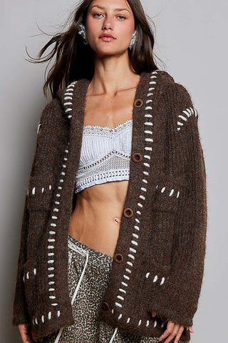 A woman wearing a brown fuzzy knit cardigan with white contrast stitching and brown buttons, layered over a white crochet crop top and patterned pants, against a plain gray studio background.