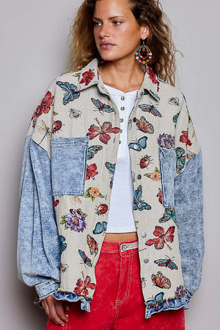 A woman wearing a patchwork button-up shirt jacket made from beige linen with colorful butterfly and floral prints, paired with blue denim sleeves, a white top, and red jeans, against a plain gray studio background.