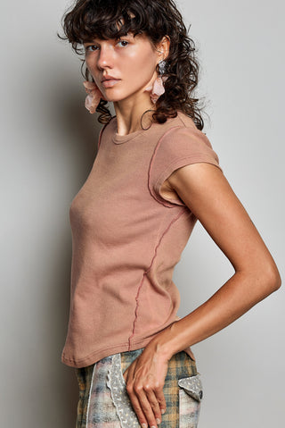 A woman wearing a fitted short-sleeve ribbed knit top in a muted dusty rose color, paired with plaid pants, photographed against a plain light gray studio background.