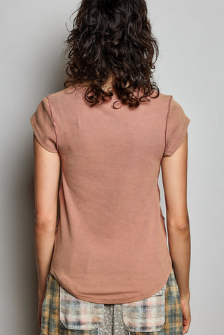 Rust-colored short-sleeve ribbed knit top shown from the back on a model with curly dark hair, paired with plaid pants against a neutral gray background.