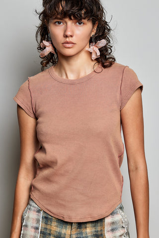 A woman wearing a short-sleeve ribbed knit top in a muted dusty rose or mauve color, paired with plaid pants, against a plain light gray studio background.