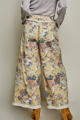 A person shown from the back wearing wide-leg cropped pants with a yellow floral print, white lace trim at the hem, and a gray long-sleeve top. The background is a plain light gray studio wall.