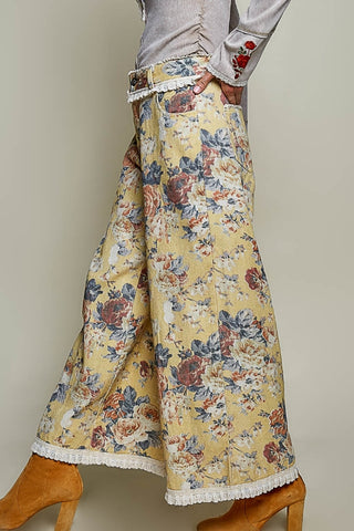 A person wearing wide-leg floral pants in a yellow base with blue, red, and beige flowers, paired with a light gray top and tan heeled boots against a plain beige studio background.
