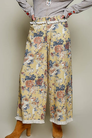 A person wearing wide-leg cropped pants with a yellow floral print, white lace trim at the waistband and hem, and tan ankle boots, shown against a plain beige studio background.