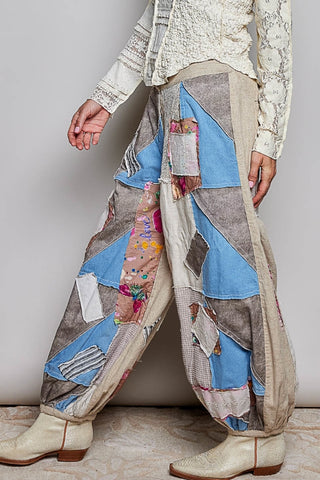 A person wearing loose, patchwork-style harem pants made of many fabric panels in beige, blue, gray, and floral prints, paired with cream lace-up boots and a cream lace-up top against a plain light gray wall.