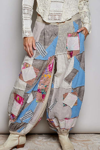 A person wearing loose, patchwork-style harem pants made from many fabric panels in beige, blue, gray, and floral prints, paired with cream lace sleeves and cream boots against a plain light wall.
