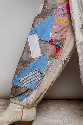 A close-up of a person's leg wearing a pair of heavily patched, distressed patchwork trousers in beige, blue, and brown, paired with a textured cream boot.