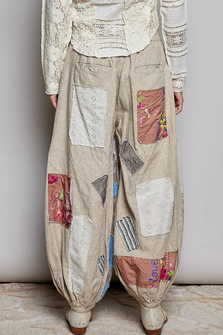 A person wearing loose beige patchwork harem-style pants with multiple fabric panels, shown from the torso down against a plain light gray wall.