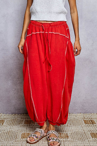 A person wearing loose, voluminous red harem-style pants with white contrast stitching and a drawstring waist, paired with a white ribbed top and patterned sandals, standing against a textured gray wall.