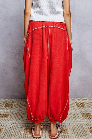 A person wearing loose, balloon-style red harem pants with white contrast stitching, paired with a white sleeveless top and sandals, standing against a textured gray wall on a woven rug.