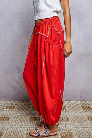A person wearing bright red harem-style pants with white contrast stitching, paired with a white textured top and patterned sandals, standing against a gray wall and woven rug.