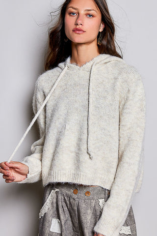 A woman wearing a fuzzy knit cropped hoodie in a light cream or off-white color, paired with gray distressed pants, against a plain light gray studio background.