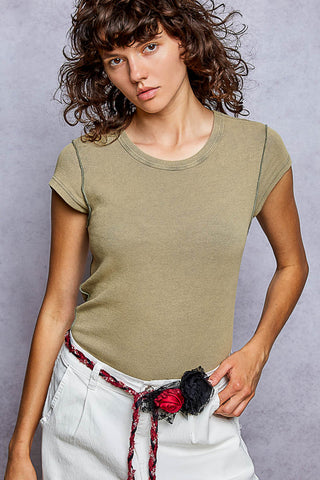 A woman wearing a fitted short-sleeve crewneck t-shirt in olive green, paired with white pants and a decorative red and black belt, posed against a textured gray studio background.