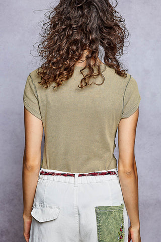 Back view of a person with long curly brown hair wearing a fitted short-sleeve olive green ribbed t-shirt and white patchwork pants against a textured gray studio background.