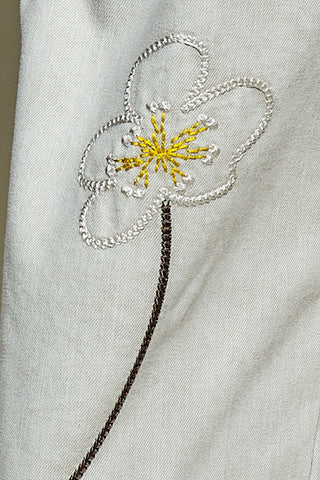 Close-up of a white fabric surface featuring an embroidered flower motif with a yellow center, silver beaded outlines, and a dark brown stem.