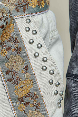 Close-up of a light gray denim garment featuring a wide gray fabric band with yellow and brown floral embroidery, silver metal studs, and a denim pocket.