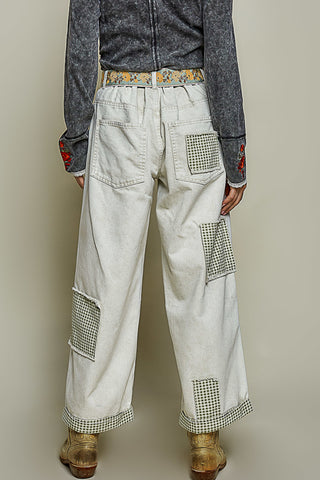 A person shown from the back wearing off-white wide-leg patchwork jeans with green-and-white gingham pocket and patch details, a gray textured top, and brown boots against a plain beige studio background.