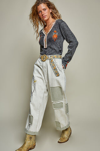 A model wearing a dark gray long-sleeve western-style top with a decorative orange graphic on the chest, paired with off-white patchwork wide-leg jeans and tan cowboy boots, posed against a solid light gray studio background.