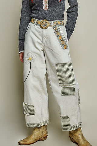 A person wearing wide-leg off-white western-style pants with embroidered floral details, checkered patch pockets, and a decorative embroidered belt, paired with a dark gray long-sleeve top and tan cowboy boots against a plain beige studio background.