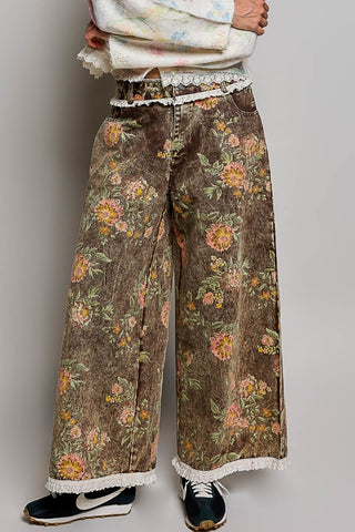 A person wearing wide-leg brown jeans with a vintage floral print, featuring white lace trim at the waist and hem, paired with black sneakers against a neutral gray background.