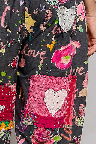 Close-up of a pair of black patchwork pants featuring a colorful floral print, hand-stitched hearts, and a prominent pink patchwork pocket with a white heart applique.