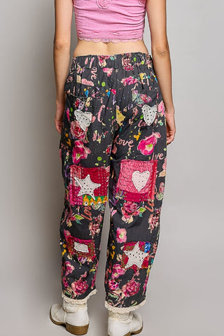 A person shown from the back wearing loose black patchwork pants with colorful floral prints, pink quilted pockets, and white lace-up boots against a light gray studio background.