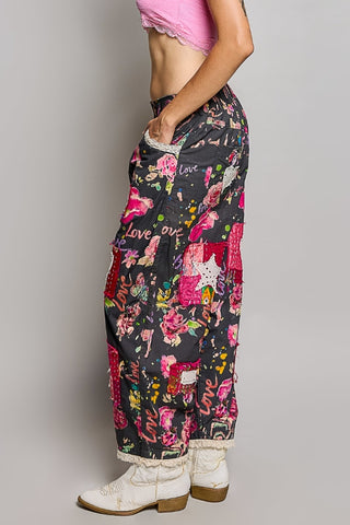 A person shown from the waist down wearing loose black patchwork pants covered in pink, red, and white floral and western-style prints, with white boots and a pink cropped top against a light gray studio background.