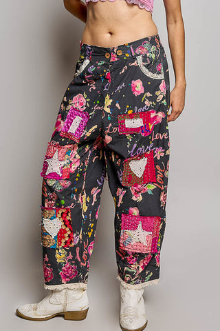 A person wearing loose black pants covered in a colorful floral and patchwork print with pink, red, yellow, and white motifs, paired with white cowboy boots against a light gray studio background.