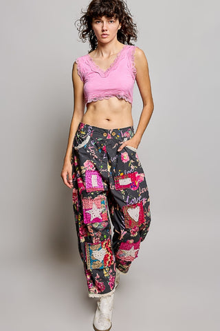A model wearing a pink ruffle-trimmed crop top and high-waisted, wide-leg patchwork-print pants with white boots, posed against a solid gray studio background.