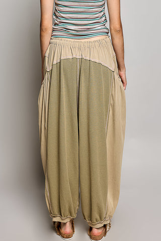A person wearing loose, baggy harem-style pants in a muted olive green and beige color-blocked design, paired with a striped tank top and sandals, shown from the back against a neutral gray studio background.