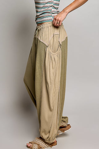 A person wearing loose, draped harem-style pants in beige and olive green, paired with a striped top and woven sandals, shown against a plain light gray studio background.