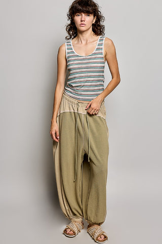 A model stands against a light gray studio background wearing a sleeveless tank top with thin multicolor horizontal stripes and loose, wide-leg olive green pants with a drawstring waist.