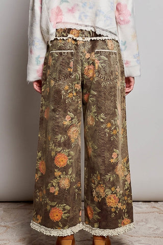 A person shown from the back wearing wide-leg cropped pants with a brown floral print, lace trim at the waistband and hem, and brown heels against a plain light gray wall.