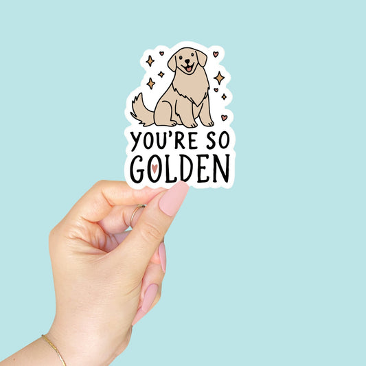 Hand holding a sticker with a dog illustration and text 'You're So Golden' against a light blue background